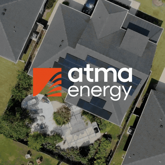 An aerial view of a gray roof with solar panels and the ATMA energy logo, situated on a residential property with green lawns and trees.